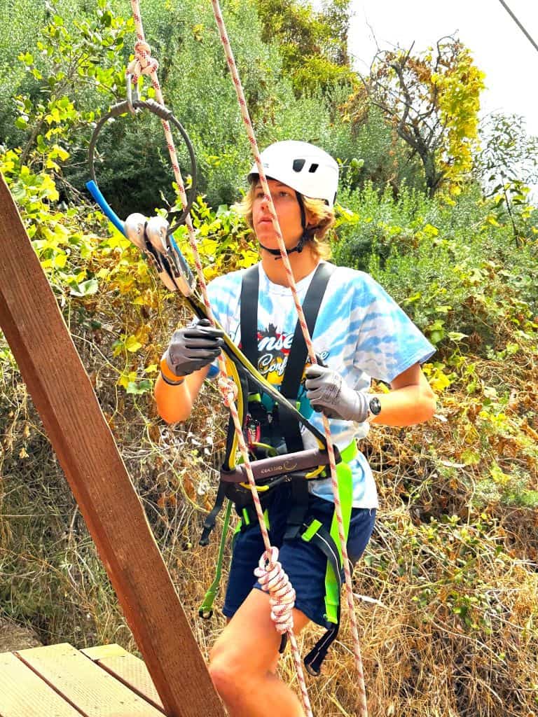 A teenaged boy wearing a helmet and harness, climbing a wooden obstacle in a forested area on Catalina Island.