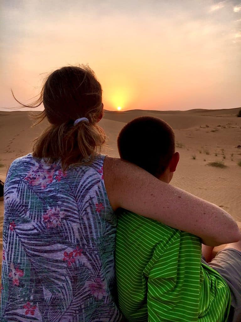 Mother and son on desert sand looking toward sunset.