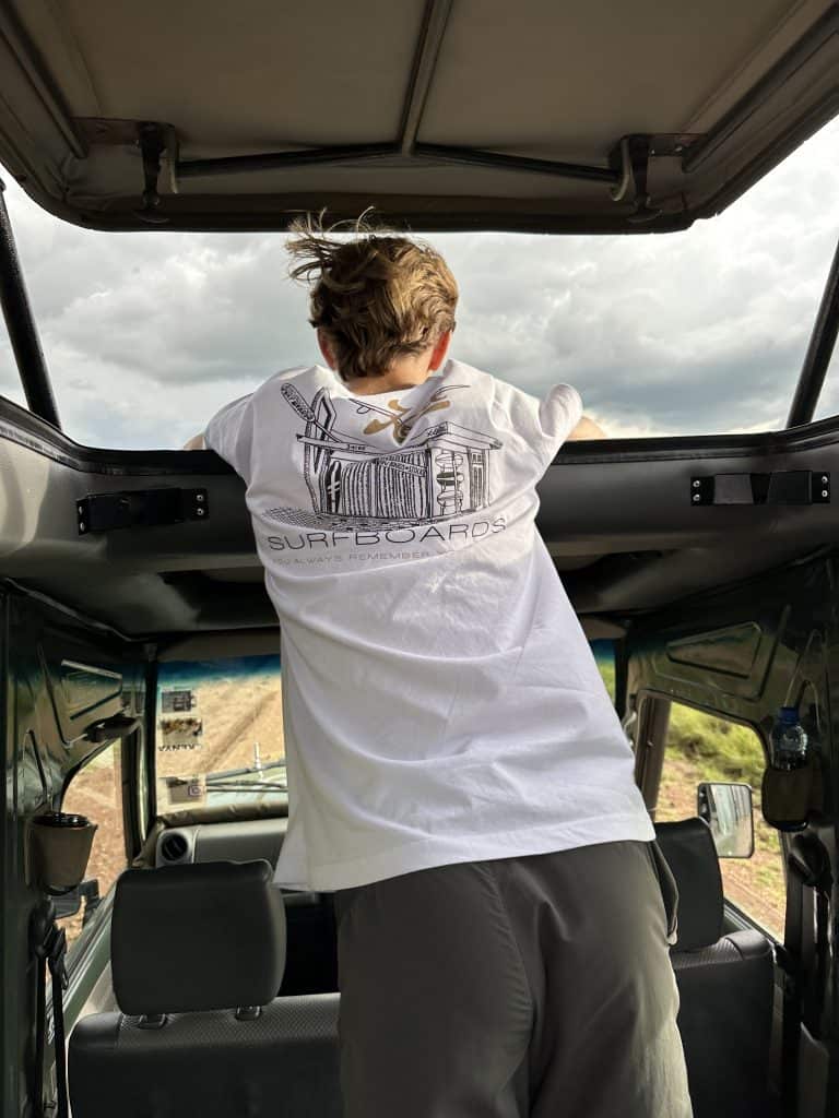 Open-air safari jeep on dirt road, with young man standing, looking for wildlife under cloudy sky.