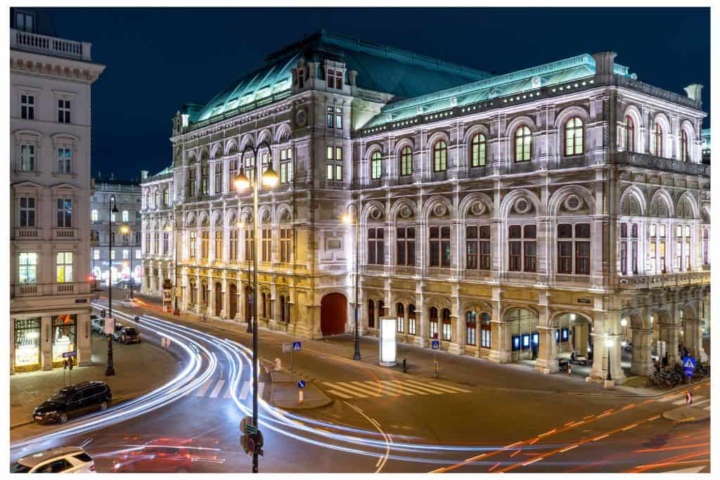 Night time scene of Vienna's Opera House, lit up with lights, as car headlights move in front of it.