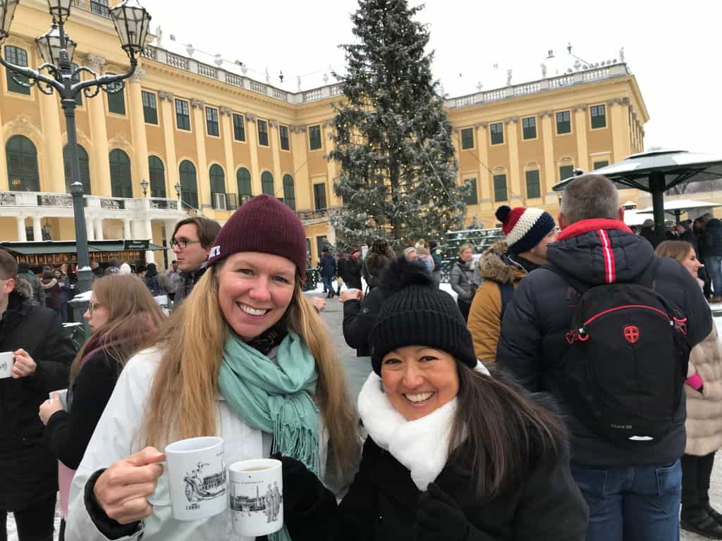 Two middle-aged women in front of large decorated tree, each holding a mug with a warm beverage.