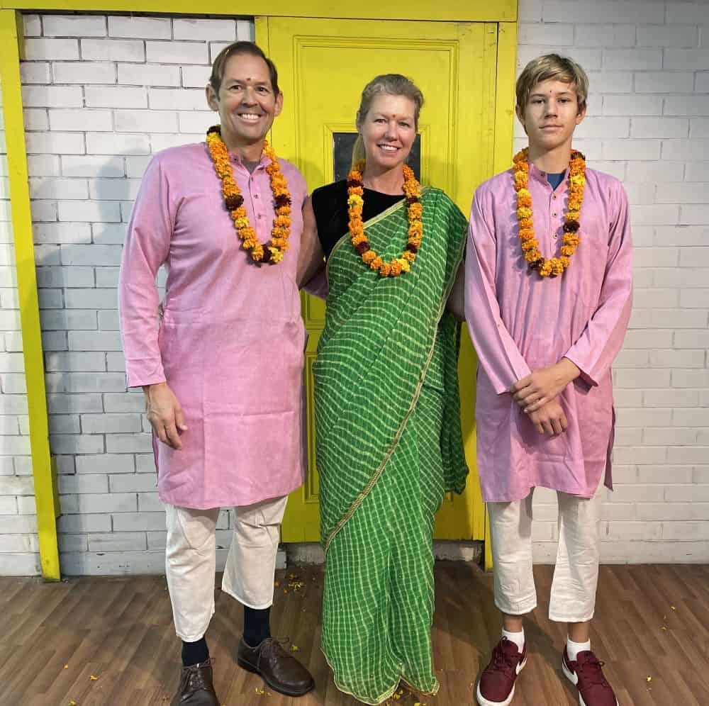 Three people, dressed in traditional Indian clothing with flowered necklaces, pose in front of a white wall and yellow door.