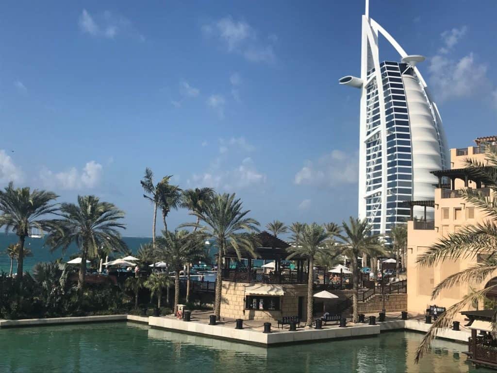 A tall white building next to the sea and a small marina with palm trees nearby.