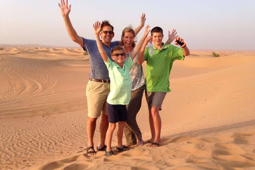 A happy family poses on the sandy desert with their arms in the air.