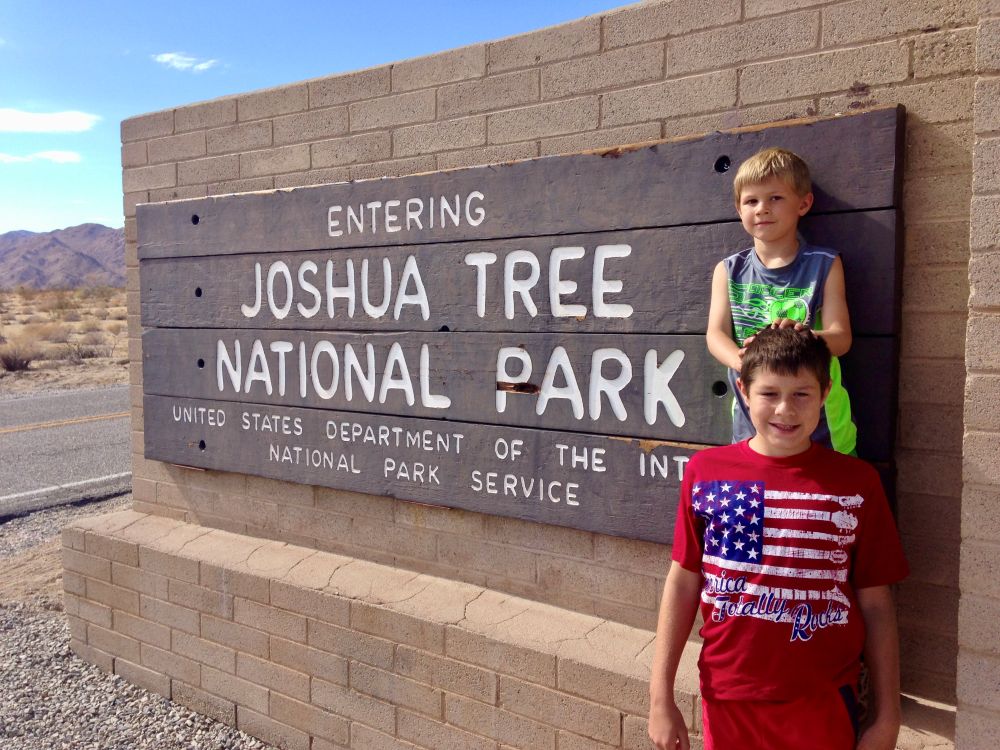 Two young boys stand before a large entrance sign that reads "Entering Joshua Tree National Park."