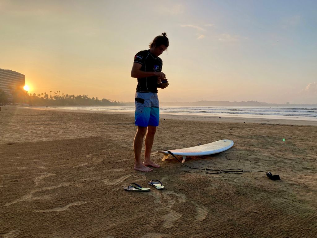 Young man standing on a beach, next to a surfboard. The sun sets behind him.