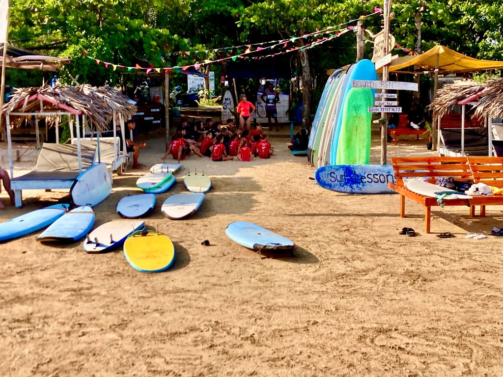 A surf shop in Sri Lanka, with a group of beginner surfers in a lesson. They are sitting on the sand and colorful surfboards are around them.