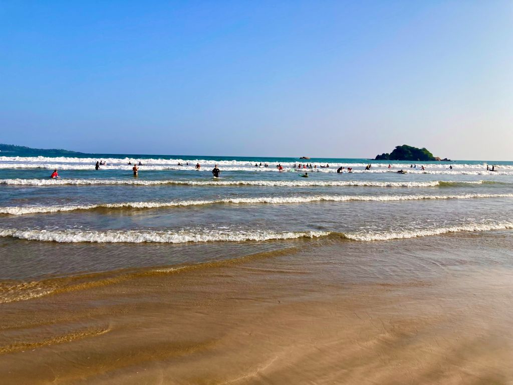 A wide sandy beach with dozens of surfers amidst several small waves in front of a rocky island in Sri Lanka.