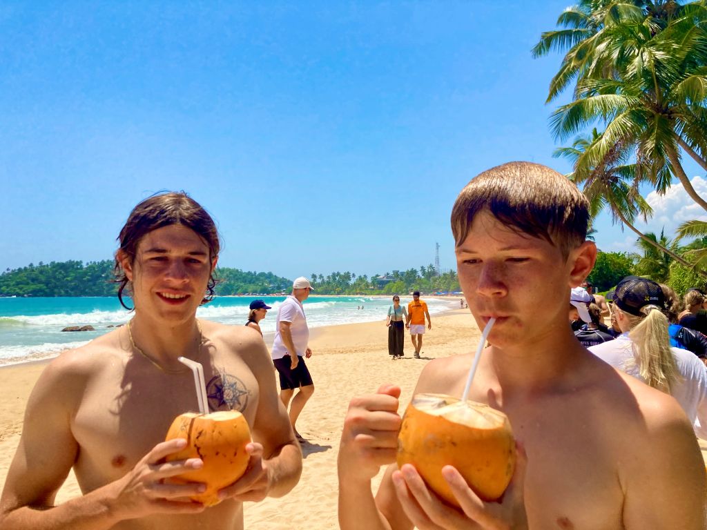 Two boys on a beach, drinking coconut water through a straw in Sri Lanka