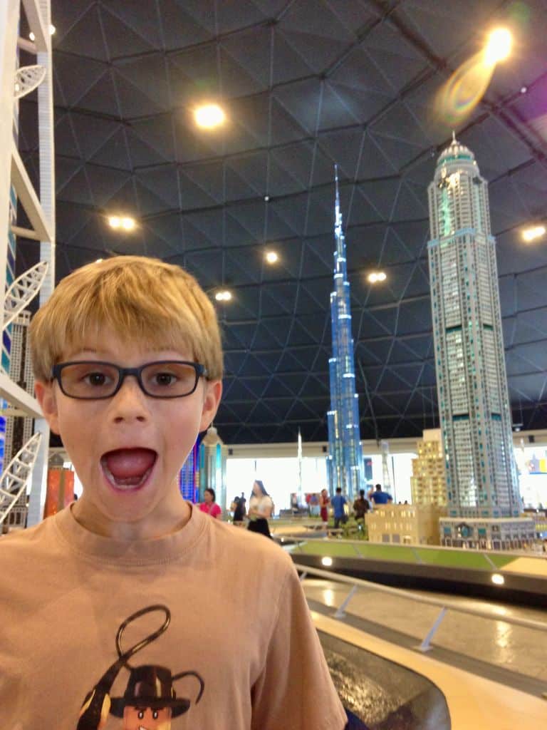 Young boy with an open mouth as if in awe, stands in a large room with huge Lego buildings behind him.