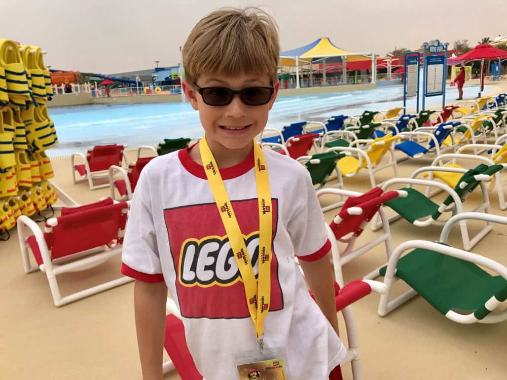 Young boy with a Lego shirt on stands in front of a large pool and many lounge chairs at Legoland Dubai's waterpark.