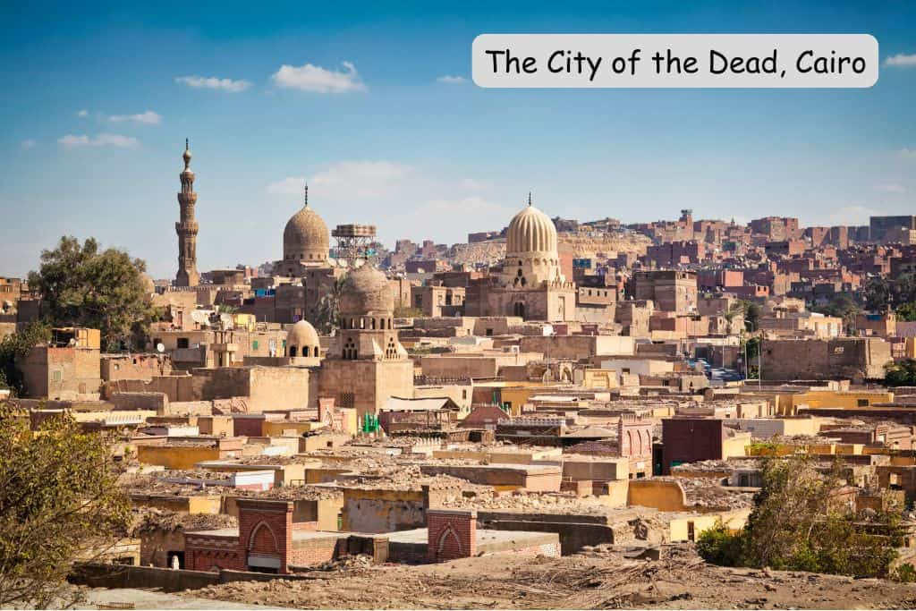 "The City of the Dead, Cairo" is written in the top right corner of an image of hundreds of stone brick buildings layered upon one another, rising up a hill. Several narrow domes on top of mosques are also seen, as well as one tall minaret.