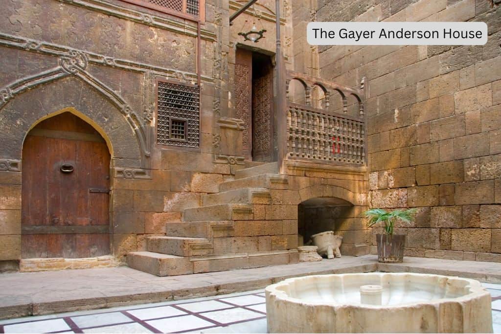 A courtyard with white tile flooring and a marble fountain in the middle. A solid stone-brick building is in the background, with a pointed arch entryway, several stone stairs leading to a wooden doorway, and decorated balcony in front. The words "The Gayer Anderson House" are overlaid on the image.