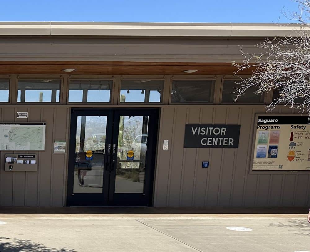The entrance to the visitor center at Saguaro National Park. The words "Visitor Center," and "Saguaro, Progams, Safety" are written on a sign next to the door.