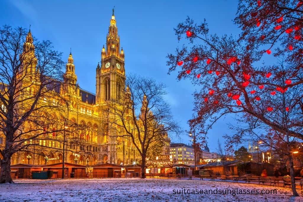 Lit majestic building in Vienna, decorated for Christmas. Snow and a tree are in the foreground.