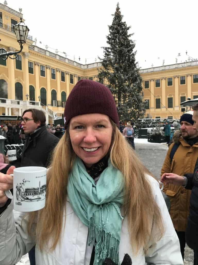 Woman with long hair and a maroon hat on is holding a coffee mug in front of a decorated Christmas tree outside a palace in Vienna.