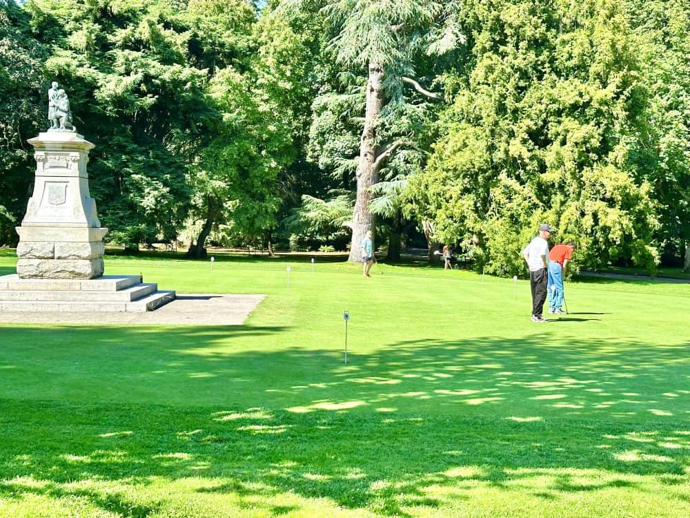 Two men with golf clubs stand on a putting green in Beacon Hill Park in Victoria, BC. The putting green has bright green grass with small stakes marking the holes. There is also a memorial statue on a pedestal in the middle of the putting green.