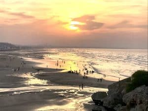 A long beach at sunset with many people wading in the shallow water near Muscat, Oman.
