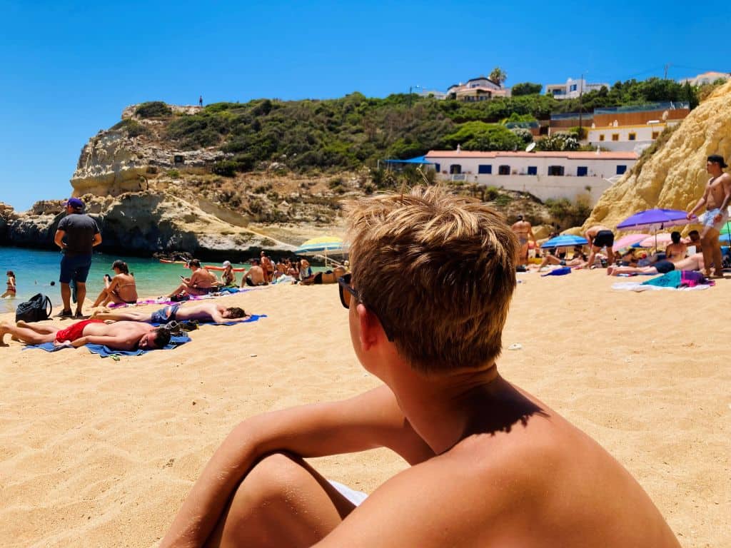 A teenaged boy sits on a sandy beach next to a rocky cliff in Portugal on a sunny day.