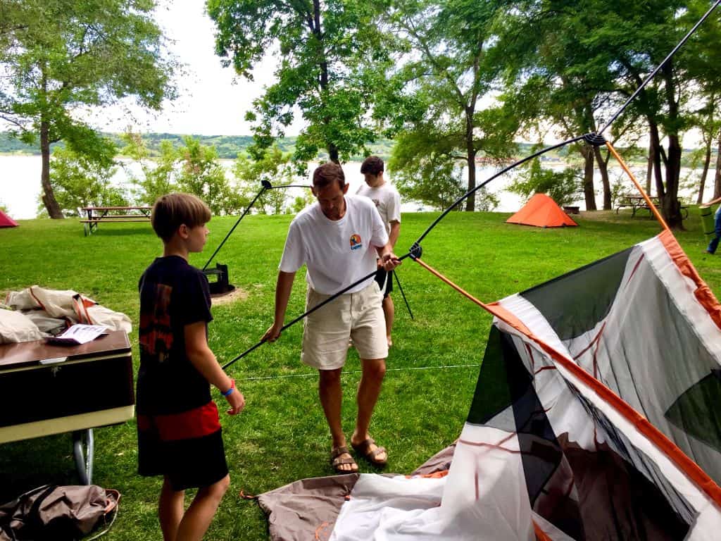 Family setting up a camping tent at a riverside campsite, with a man assembling tent poles while two boys help on a grassy area surrounded by trees 