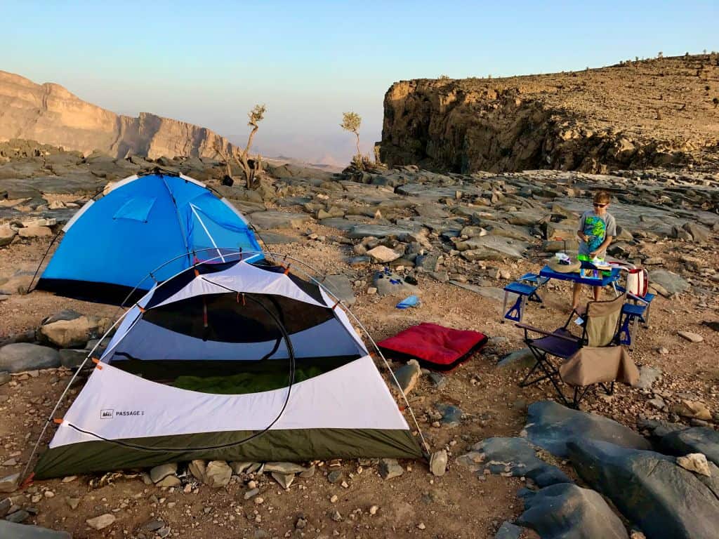 Family campsite set up on rocky desert terrain with two tents, camping chairs, and a child preparing food at a picnic table near a canyon at sunset.