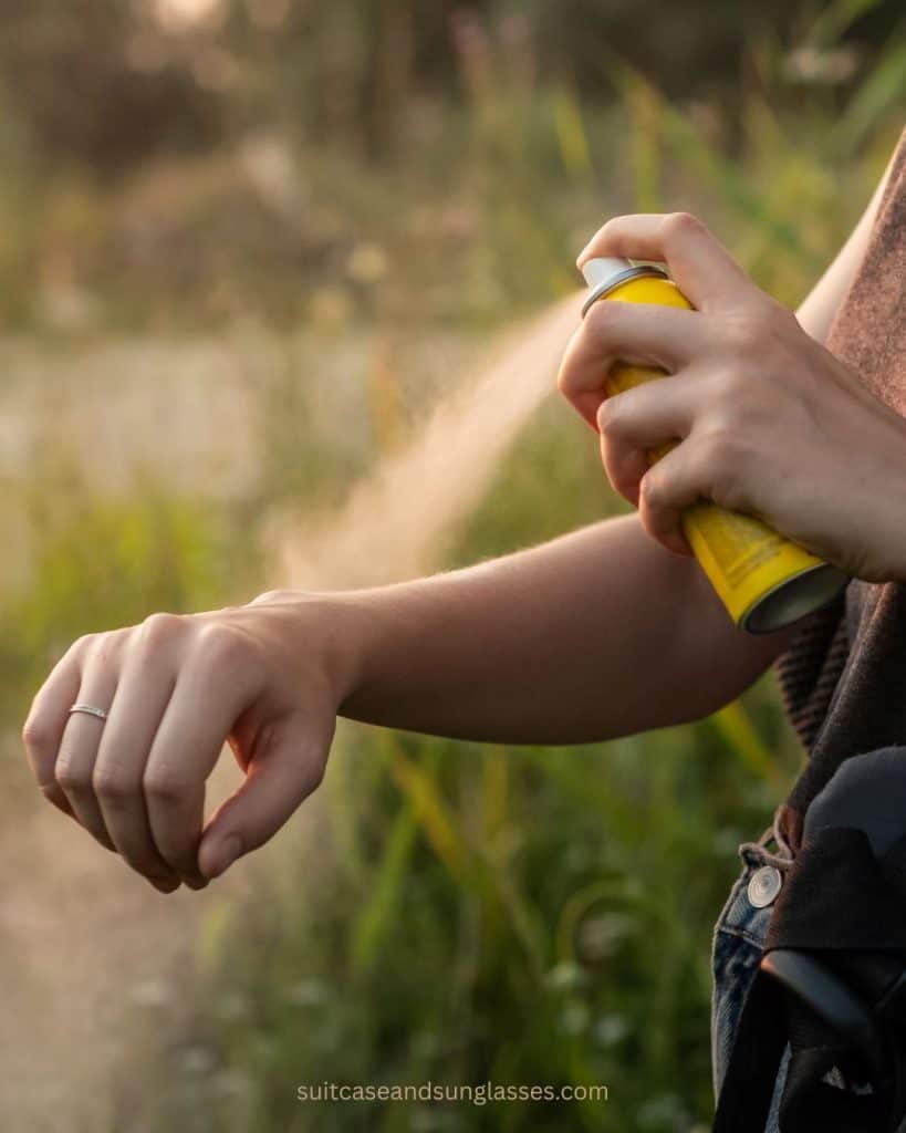 Applying mosquito repellent spray to arm while outdoors on a grassy trail at sunset during camping trip 