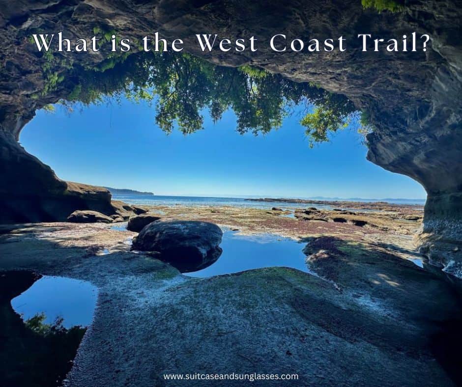 A scenic view toward the ocean through the mouth of a cave with green bushes hanging over the entrance. Large rocks and boulders are at the entrance of the cave, and the coastline and islands are seen in the distance. "What is the West Coast Trail?" is overlaid text.