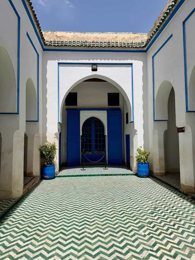 Intricate green and white geometric tiled floor in a courtyard of Bahia Palace in Marrakech, Morocco, surrounded by white archways and blue doors.
