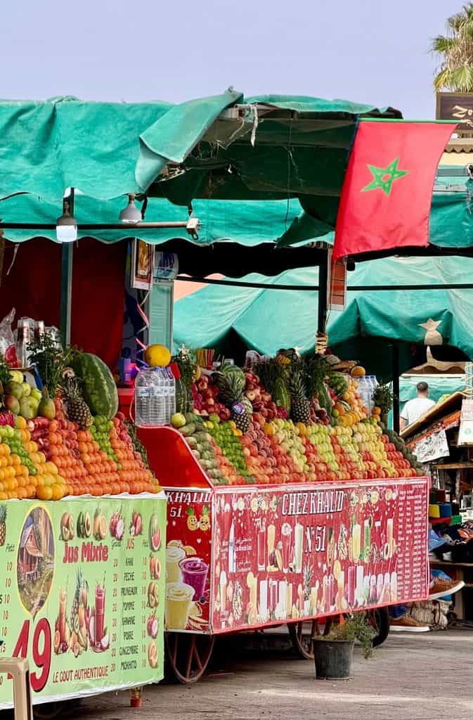 Colorful fresh fruit and juice stalls at Jemaa el-Fnaa square in Marrakech, Morocco, with oranges, pineapples, watermelons, and a Moroccan flag overhead.