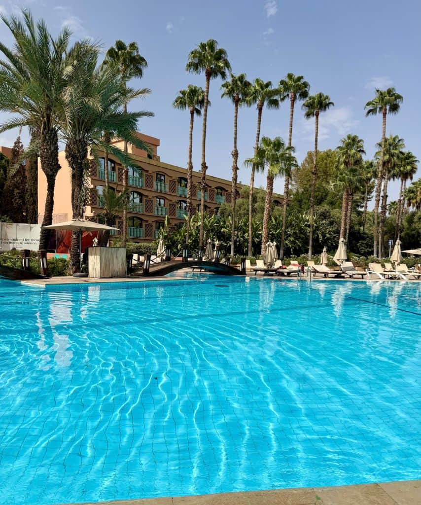 Large turquoise swimming pool surrounded by palm trees, lounge chairs, and hotel balconies at Le Méridien N'Fis in Marrakech, Morocco.