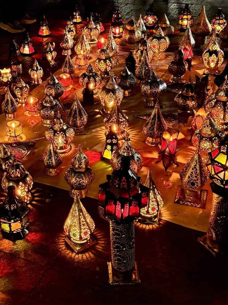 Glowing Moroccan metal lanterns with intricate cutout patterns and colorful glass displayed for sale in the Marrakech medina at night.