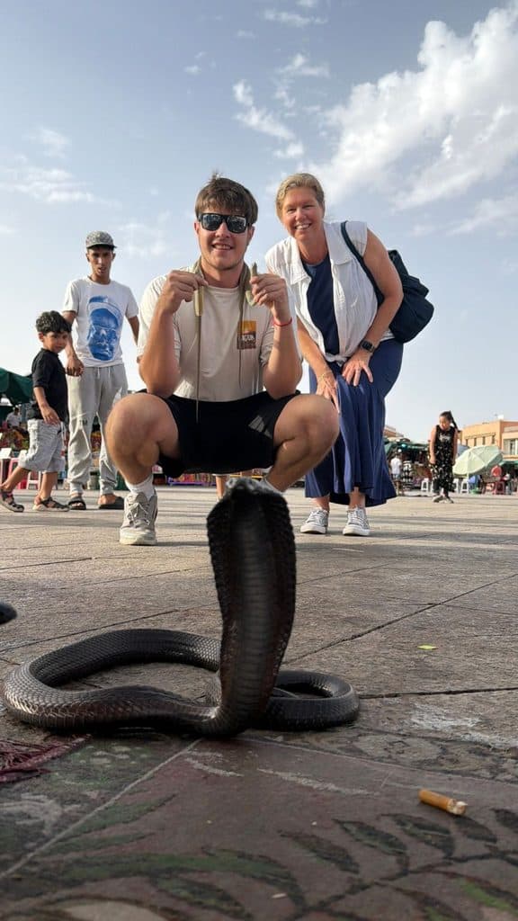 Large cobra with its hood raised in the foreground as two travelers pose behind it at Jemaa el-Fnaa square in Marrakech, Morocco.