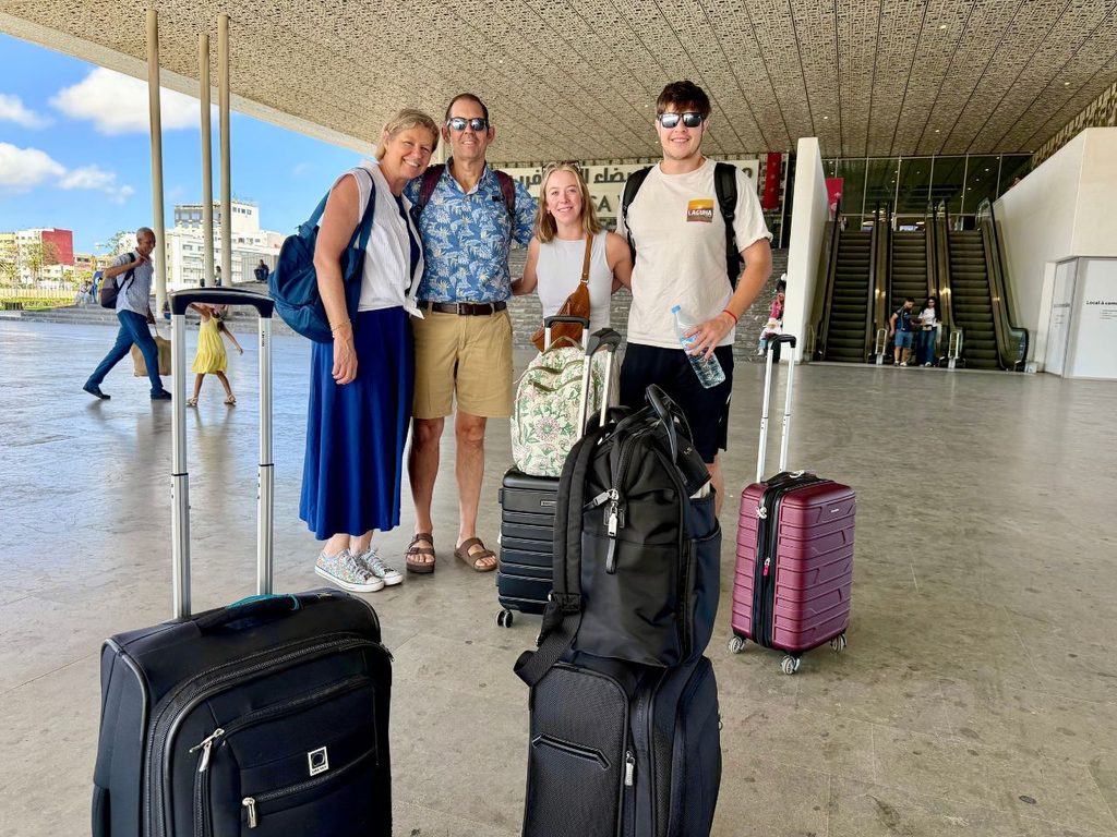 Four travelers with small rolling suitcases and backpacks outside a train station in Morocco.