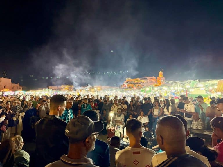 Crowds gathered around performers and brightly lit market stalls at Jemaa el-Fnaa square in Marrakech, Morocco, at night.