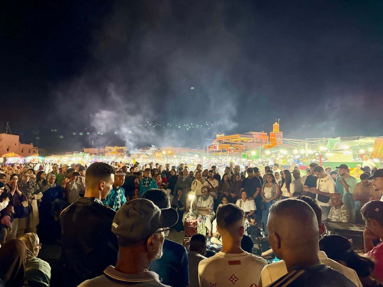 Crowds gathered around performers and brightly lit market stalls at Jemaa el-Fnaa square in Marrakech, Morocco, at night.